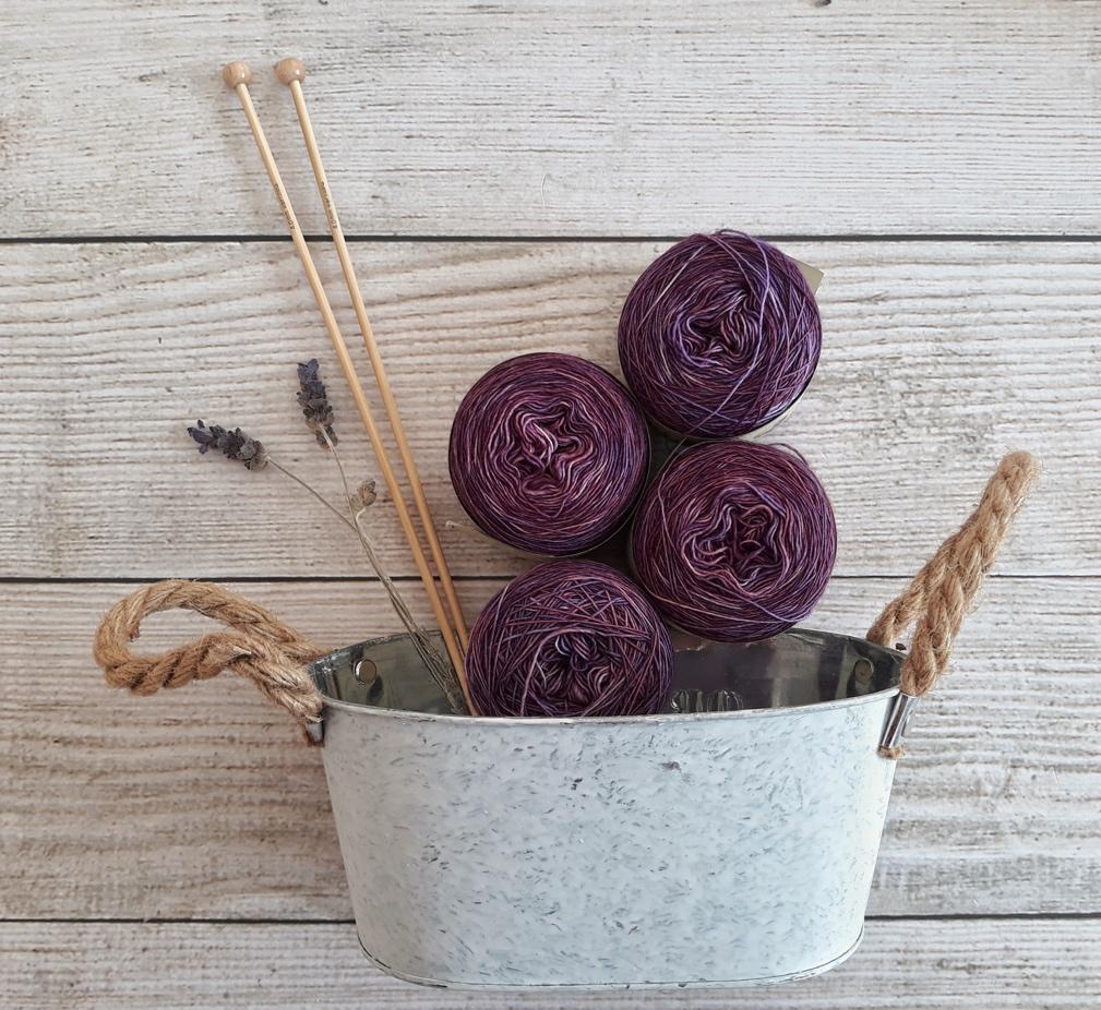 Four balls of purple yarn along with wooden knitting needles and a spray of greenery arranged in a pail on a wooden background