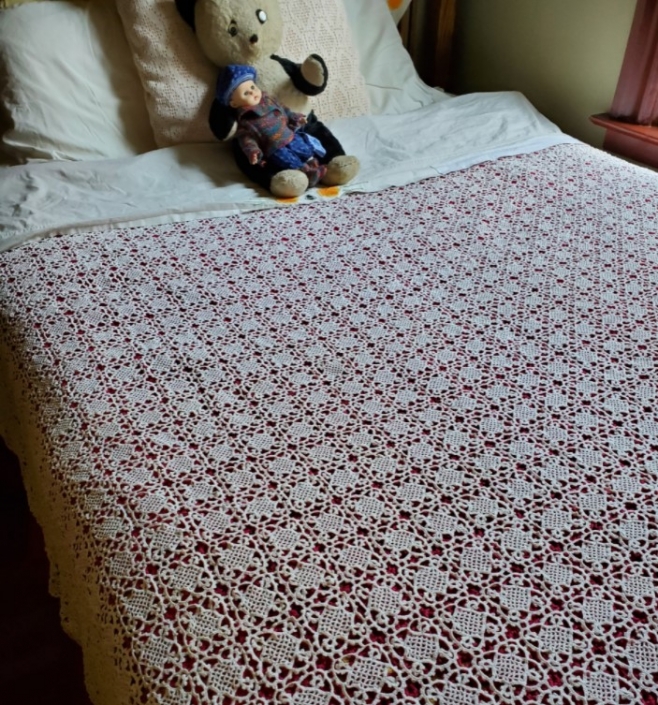 coverlet of assembled lacy crocheted squares on a large bed with folded over top sheet that is embroidered with a poppy motif, with a stuffed panda bear and a baby doll lying on the pillows