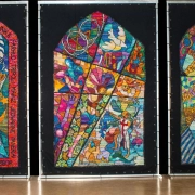 Kirk Dunn standing in front of three multicolored knit panels depicting elements of the Jewish, Christian, and Islamic faiths