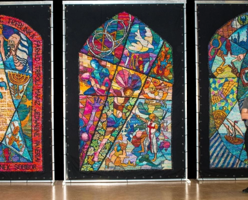Kirk Dunn standing in front of three multicolored knit panels depicting elements of the Jewish, Christian, and Islamic faiths